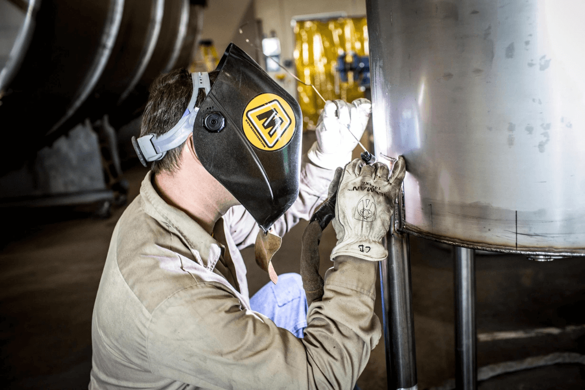 An MXD Process welder wearing a fire safety mask, with the MXD Process logo on it, while welding a tank cylinder tank.